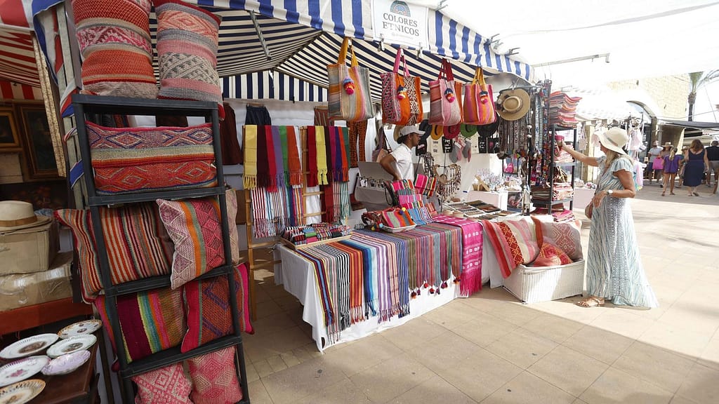 A vibrant market stall at Sotogrande Marina selling colorful handmade textiles, bags, and accessories, with a woman browsing the items.