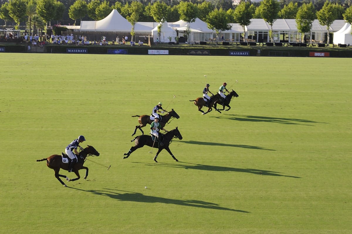 Players competing in a polo match on a green field in Sotogrande, with spectators and white tents in the background.