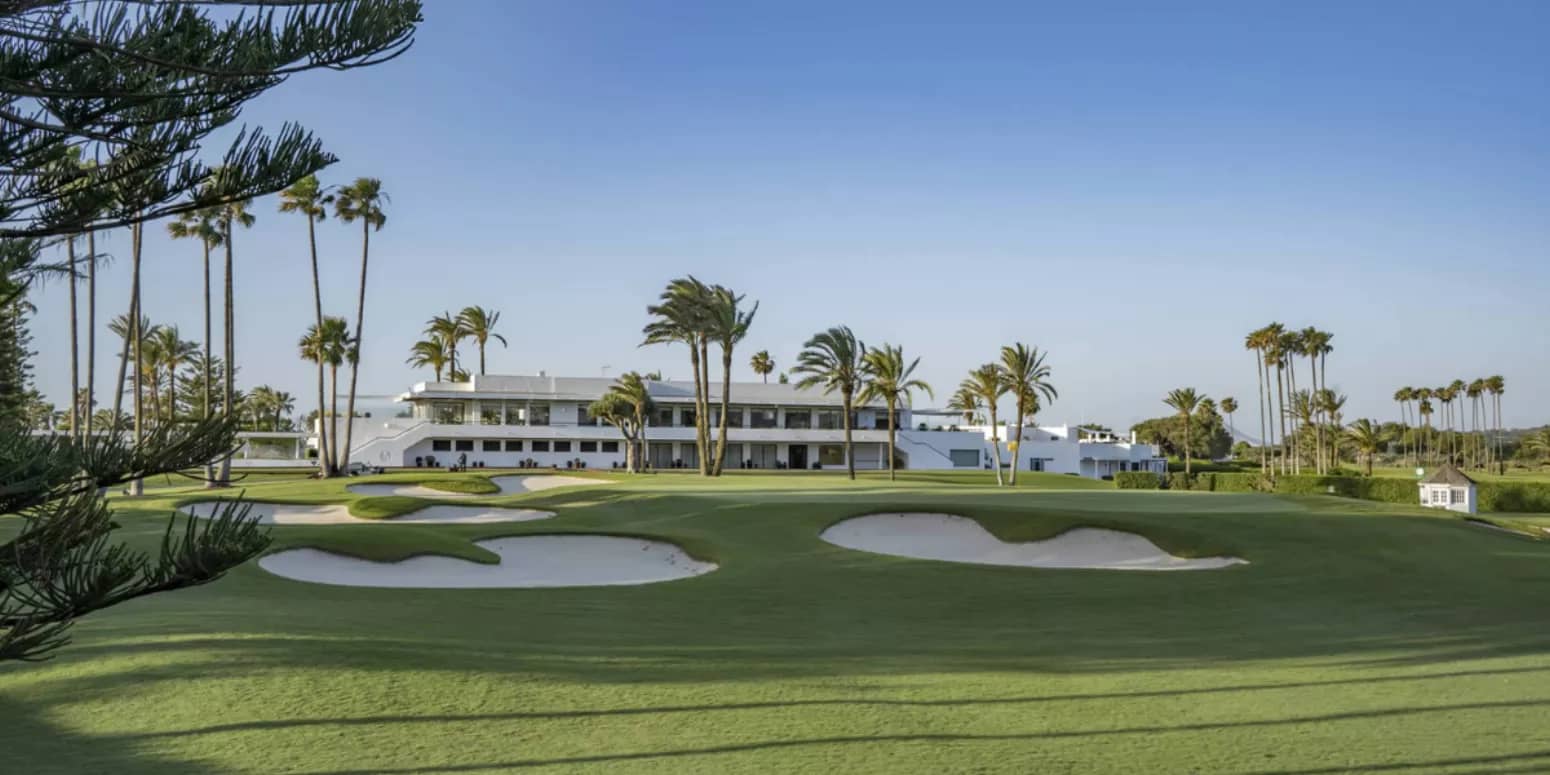 View of the lush, manicured greens of a golf course with multiple sand traps in front of the best luxury property, Real Club Sotogrande, surrounded by tall palm trees under a clear sky in Costa Del Sol, Andalucia.