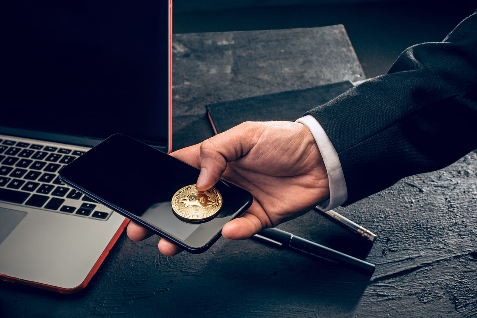 Hand holding a Bitcoin coin over a smartphone, with a laptop in the background