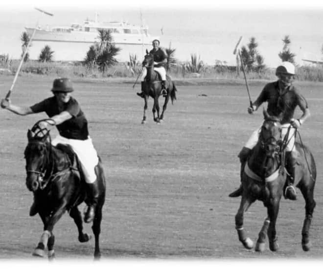 Black-and-white image of polo players on horseback in Sotogrande, with a yacht and trees visible in the background.