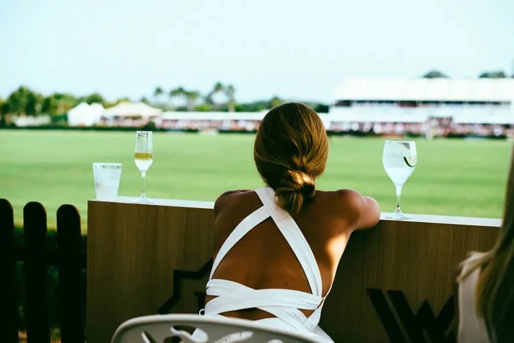 A woman in a white dress watching a polo match in Sotogrande, with drinks on the table and a green field in the background.